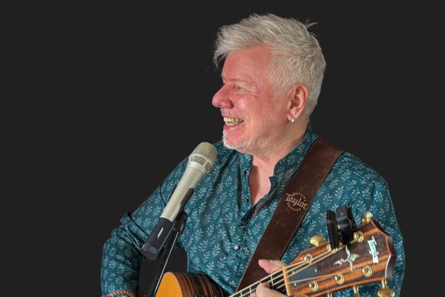 Musician Gordon Mark Webber smiling and playing an acoustic guitar during a live music event at Indian Essence in Orpington