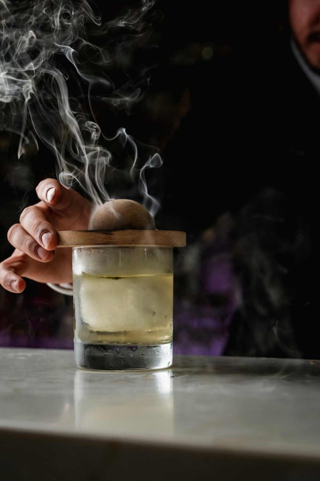 A bartender lifting a wooden smoking cloche to reveal a freshly prepared smoked cocktail over a large ice cube at Indian Essence, Petts Wood.