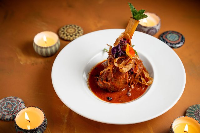Spiced lamb shank curry garnished with edible flowers, surrounded by glowing Diwali diyas on a warm copper surface.
