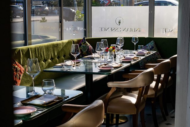 A sunlit, elegantly set dining table with wine glasses, green velvet banquette seating, and leather chairs prepared for the wine dinner event at Indian Essence.