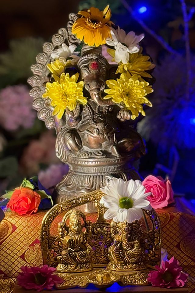 Bronze Lord Ganesha statue adorned with yellow and white flowers on a festive shrine with blue ambient lighting.