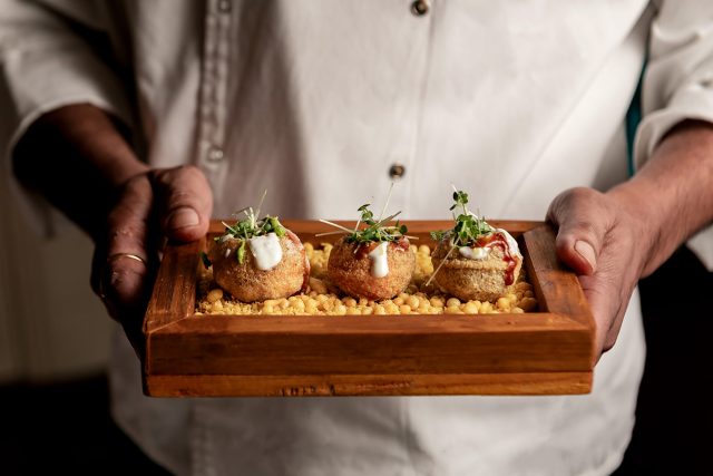 A chef holding a rustic wooden tray presenting three pieces of stuffed pani puri garnished with yogurt, chutney, and microgreens.