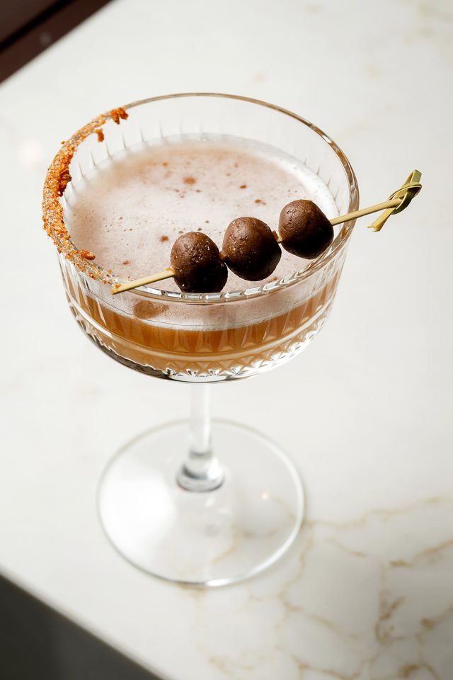 A signature cocktail in a ribbed coupe glass featuring a spiced rim, garnished with three small sweet spheres on a bamboo skewer.